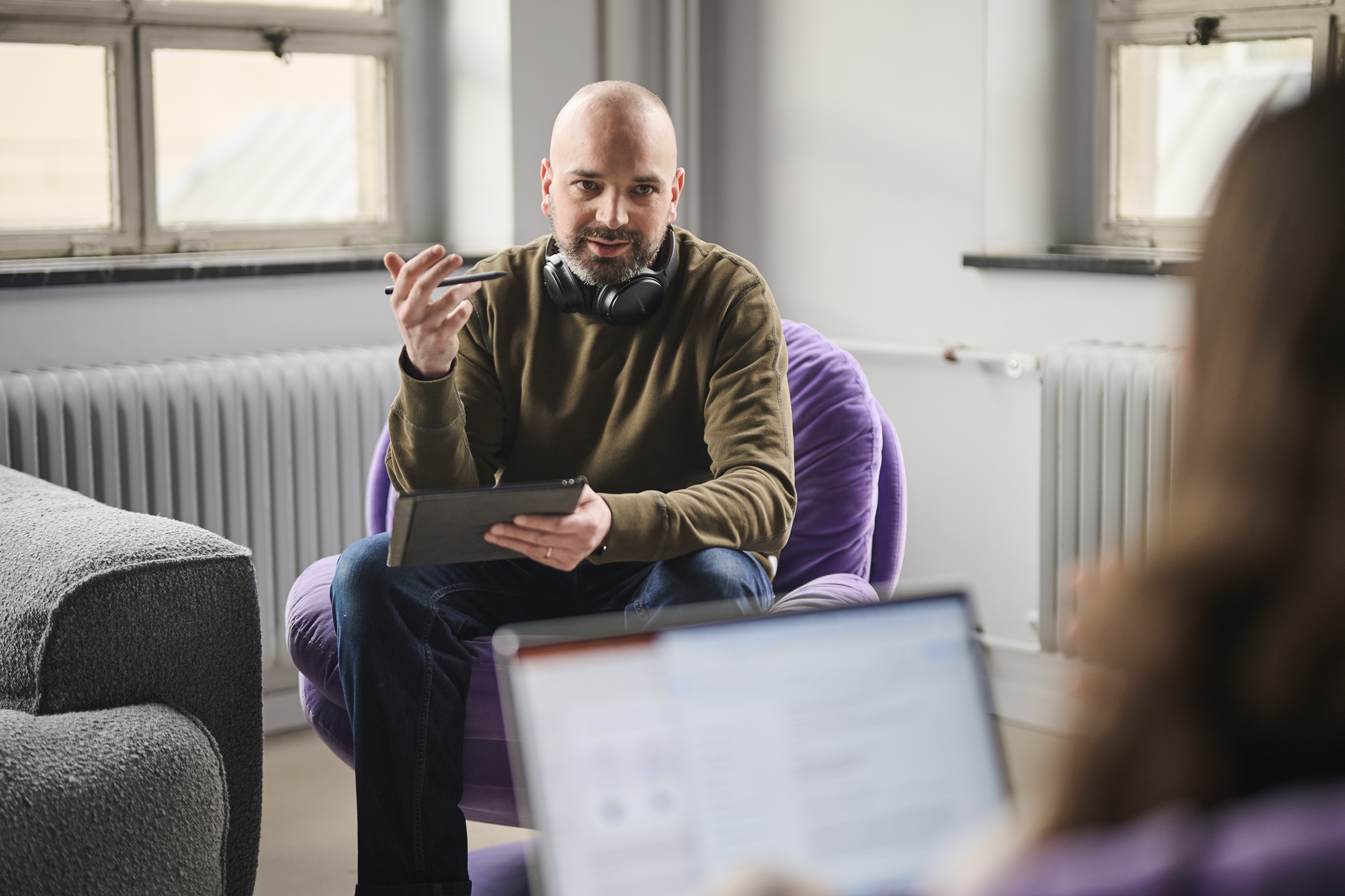 Two Colleagues, A Woman Working on her Laptop and Sitting in Front of a Man Talking About Work in The Office.